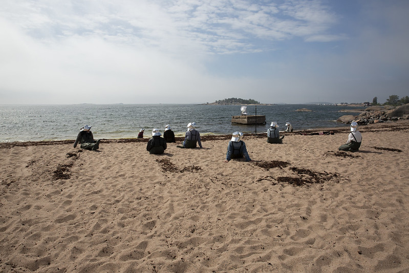 Valkoisiin hattuihin pukeutuneita ihmisiä istuu rannalla katsoen merta kohti.