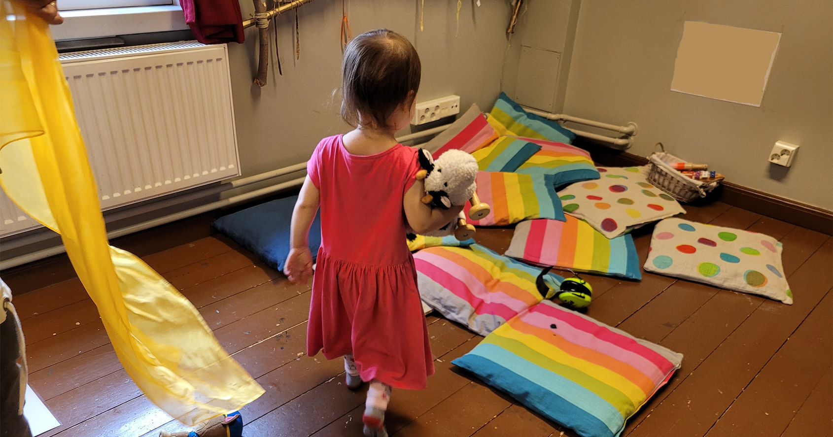 Pieni tyttö vaaleanpunaisessa mekossa, pehmolelu kädessään kulkee kohti värikästä lattiatyynykasaa.