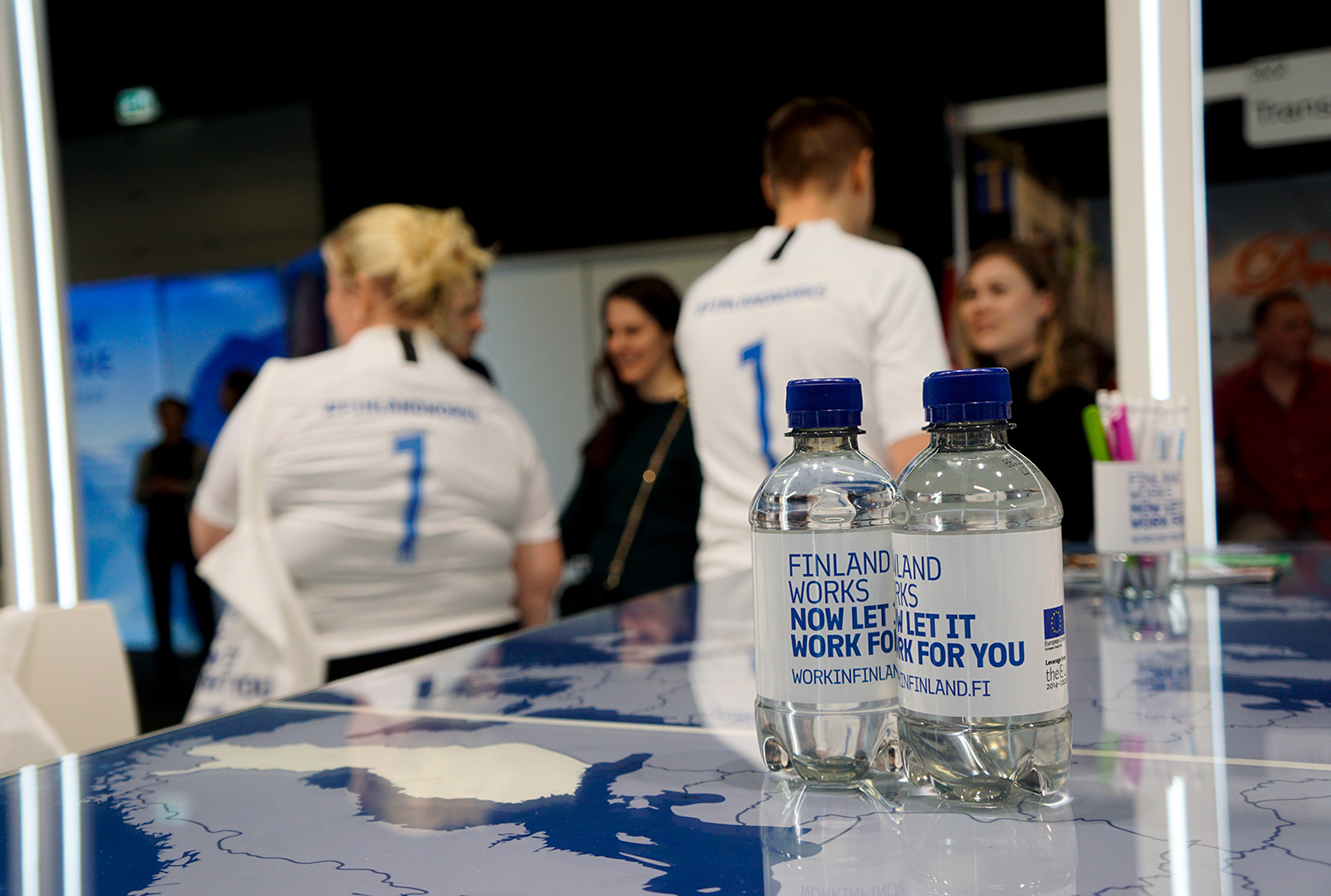 Kaksi Finland Works -etiketeillä varustettua vesipulloa messupöydällä. Taustalla ihmisiä.