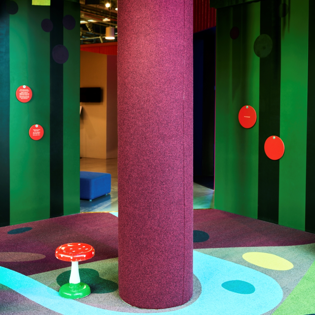 Huone, jossa on keskellä lila pylväs, värikäs matto ja vihreät seinät, joissa punaiset kurkistusluukut sekä lattialla sienituoli.