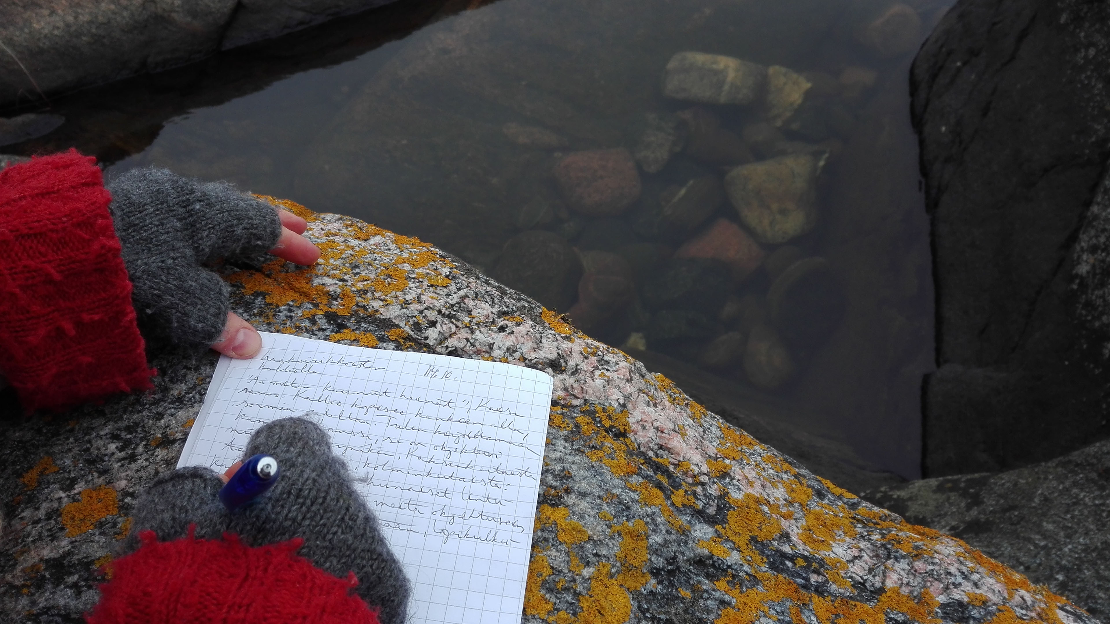 Ihmisen villakäsineisiin verhotut kädet veden äärellä, kiven päällä kirjoittamassa kynällä paperille.