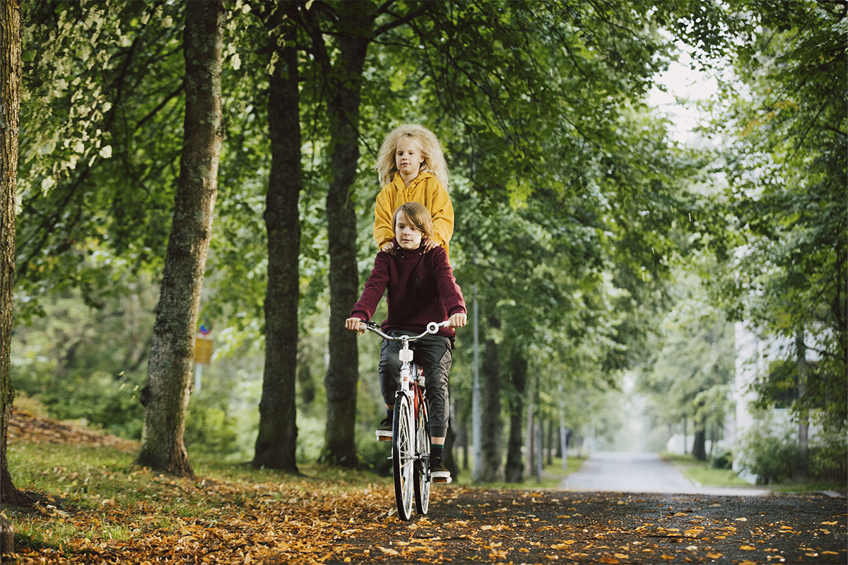 Alfred ajaa polkupyörällä ja Sihja-keiju seisoo pakkarilla. Kujaa reunustavat vehreät puut ja maassa on keltaisia lehtiä.