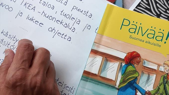 valokuva kädestä, muistilapusta ja suomenkielen oppikirjasta