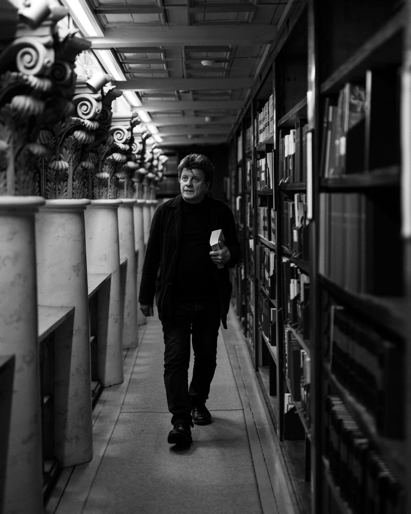 Näyttelijä Timo Torikka kävelee kirjaston käytävää pitkin kirja kädessä.